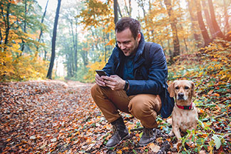 Mavi geçiş ceketli ve hava koşullarına dayanıklı erkek çizmeleri olan bir adam sonbahar ormanında bir gecekonduda diz çöküyor ve akıllı telefonunu işletiyor, yanında köpeği yatıyor.
