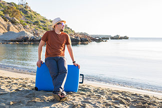 Ein junger Mann, der einen Hut trägt, sitzt am Strand auf einem blauen Herrenkoffer.