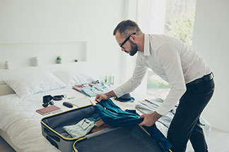 Ein Mann, der ein weißes Hemd und eine schwarze Hose trägt, packt seinen Koffer, der offen auf dem Bett liegt.