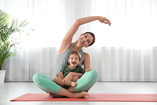En kaliteli yoga kıyafetleri: Genç bir anne, yoga matının üzerinde oturmuş kolunu başının üzerine kaldırmış, oğlu kucağında oturuyor.