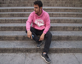 Elinde akıllı telefonu olan genç bir erkek, pembe kapüşonlu sweatshirt'üyle bir taş merdivenin üzerinde oturuyor.