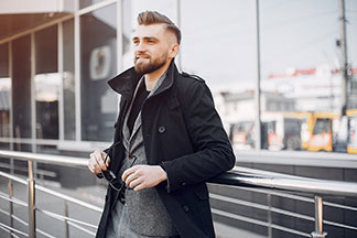 Ofis için erkek ceketi: Kısa bir siyah palto ve altında gri bir spor ceket giyen genç bir erkek, parmaklıklara yaslanmış ve güneş gözlüklerini elinde tutuyor.