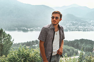 Kareli, önü açık kısa kollu gömlek giyen genç bir erkek, arka planda dağların göründüğü bir gölün önünde duruyor.