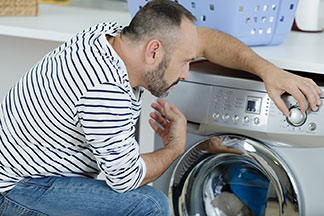 Kot pantolon ve beyaz-siyah çizgili kazak giymiş bir erkek, eşofman altını yıkamak için çamaşır makinesinin önünde diz çöküyor ve programı ayarlıyor.