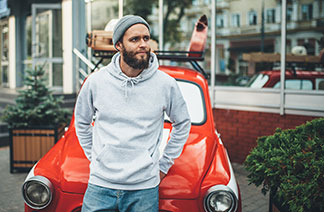 Yün şapkalı ve gri kapüşonlu sweatshirt'lü genç bir erkek, kırmızı bir arabaya rahat bir şekilde yaslanıyor.
