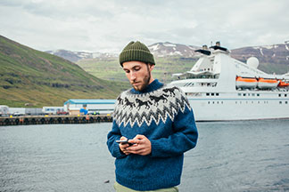 Norveç kazaklı genç, sakallı bir erkek, bir yolcu gemisinin geldiği limanın önünde duruyor ve akıllı telefonuna bakıyor.